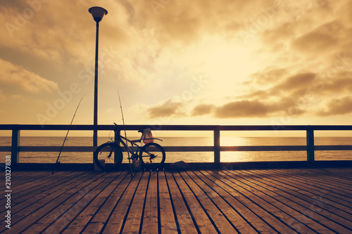  Fisherman bicycle with fishing rods on wooden bridge. Toned. Bicycle on sea bridge at sunset.  © vaitekune