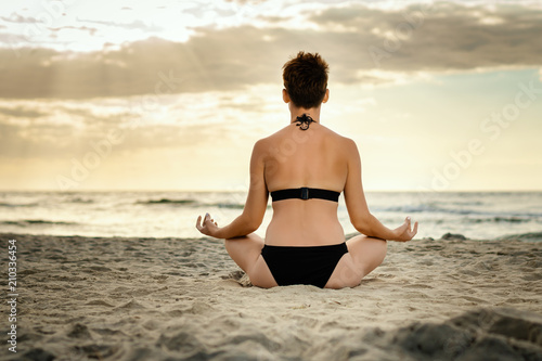 beautiful woman in a black swimsuit meditating on the beach at sunset