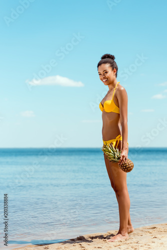 smiling african american woman in bikini holding pineapple on sandy beach