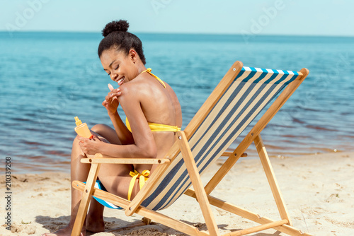 smiling african american woman applying sunscreen lotion on skin while sitting on deck chair on sandy beach © LIGHTFIELD STUDIOS