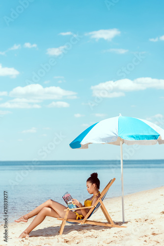 side view of young african american woman reading magazine and relaxing on deck chair under beach umbrella in front sea