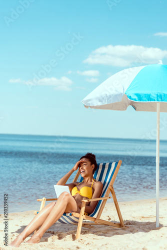 laughing african american woman relaxing on deck chair and using digital tablet under beach umbrella in front of sea