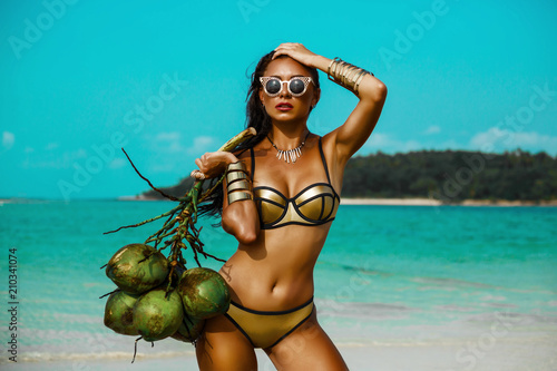 beautiful tanned girl in trendy bathing suit sunglasses stands with a bunch of coconuts in her hands on beach tropical island. Summer vacation, travel around the world, advertising swimwear new season photo