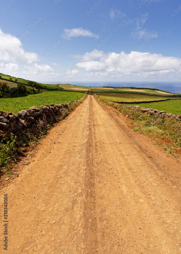 A dead-straight track leads through a hilly landscape to the sea, on the edge of natural stone walls, great depth, sky with some clouds - Location: Azores, Sao Jorge Island A dead-straight track leads through a hilly landscape to the sea, on the edge of natural stone walls, great depth, sky with some clouds - Location: Azores, Sao Jorge Island