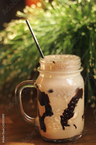 cold coffee frappe in glass jar with chocolate and foam in wooden table with fresh grass on background 