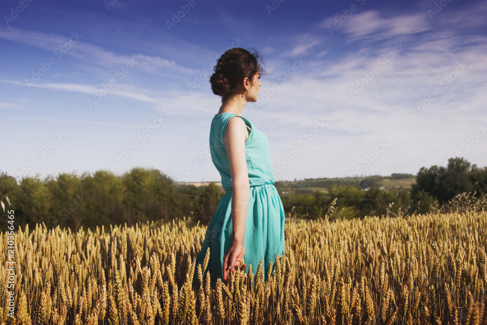 Naklejka premium A girl in a blue dress on a gold boundless wheat field looks to the sky