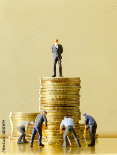 miniature business man standing on top of golden coins while looking at engineer worker team working on his money below