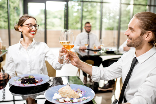 Beautiful couple clinking with wine glasses during a business lunch with delicious meals at the modern restaurant with business people on the background