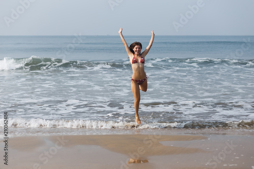 Slender young girl in a swimsuit is jumping on the sandy shore of the ocean. Happy Days. The concept of freedom, recreation, vacation. Flight of joy and fun. Reflection in water. Charming smile.