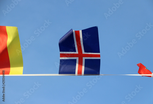 Mini fabric rail flag of Iceland in The Red Cross crosses over the white cross on the blue color hanging on the rope cloth between the flag of other countries and flick up, on blue sky background.
