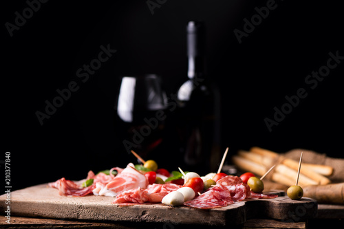 Assorted meats and cherry mozzarella cheese, on a wooden cutting board with bottle of wine and glass on background. Italian antipasti