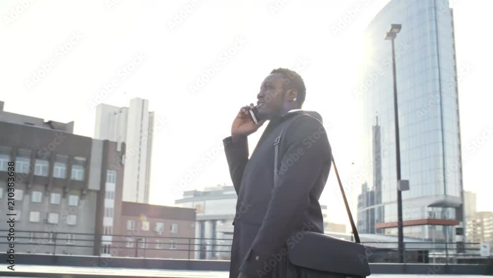 Panning shot of African American businessman standing outdoors in front of modern office building in downtown and talking on cell phone at sunny day Panning shot of African American businessman standing outdoors in front of modern office building in downtown and talking on cell phone at sunny day