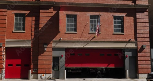 A daytime exterior establishing shot of a typical red brick fire station with the garage doors partially open in the downtown district of a large city.  	