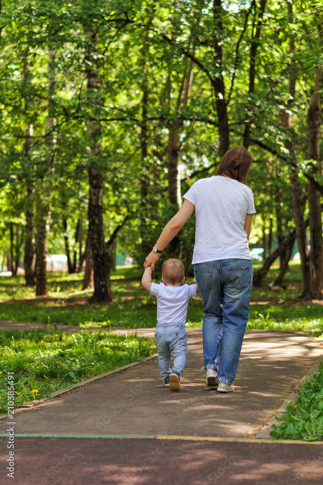 Family. Mother and son outdoors. Woman holds child by hand. Baby learns to walk. Shooting from the back. People are unrecognizable Family. Mother and son outdoors. Woman holds child by hand. Baby learns to walk. Shooting from the back. People are unrecognizable