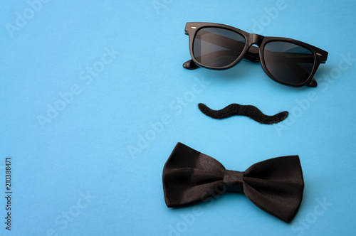 Father day and male hipster fashion concept with minimalist image of a pair of square sunglasses, black bowtie and a fake moustache on colorful blue background with copy space