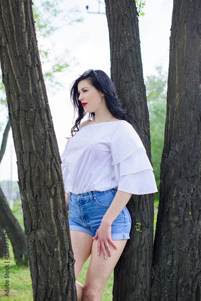 Portrait of pretty black hair young woman in the style black sunglasses on the background of city park and garden. Girl wearing in white romantic blouse and short jeans with high waist. Portrait of pretty black hair young woman in the style black sunglasses on the background of city park and garden. Girl wearing in white romantic blouse and short jeans with high waist.