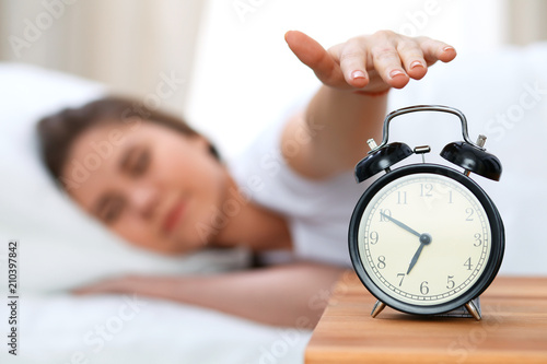 Sleepy young woman stretching hand to ringing alarm willing turn it off. Early wake up, not getting enough sleep concept