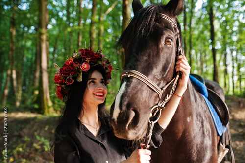Mystical girl in wreath wear in black with horse in wood.