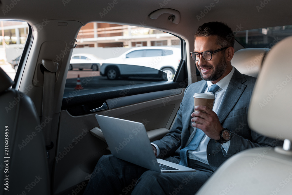 smiling businessman in eyeglasses with coffee to go using laptop on backseat in car