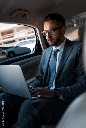 businessman in eyeglasses using laptop on backseat in car