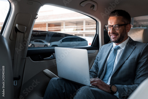 smiling businessman in eyeglasses using laptop on backseat in car © LIGHTFIELD STUDIOS