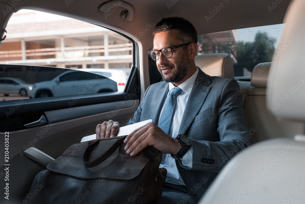 businessman in suit putting papers into bag while sitting on backseat in car