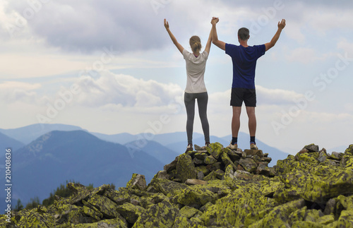 Back view of young couple, athletic boy and slim girl standing with raised arms on rocky mountain top enjoying breathtaking summer mountain view. Tourism, success and healthy lifestyle concept.