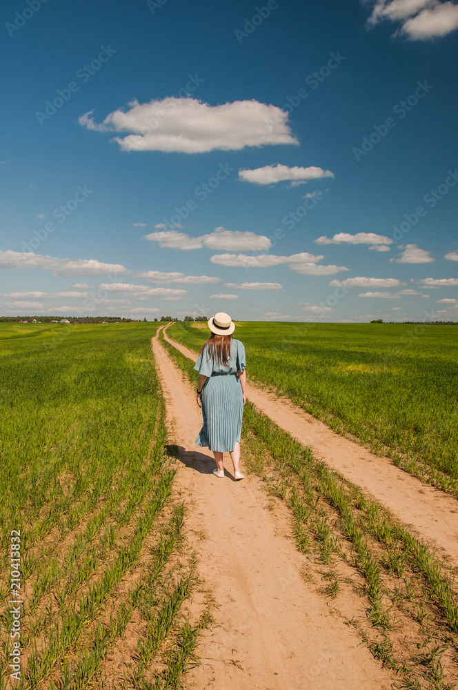 Beautiful young romantic girl outdoor in a blue dress and a straw hat with long hair enjoying nature, walking, spinning and dancing on the green field in the village