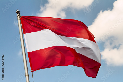 Fotografie Flag of Austria waving in the wind on flagpole against the sky with clouds on su