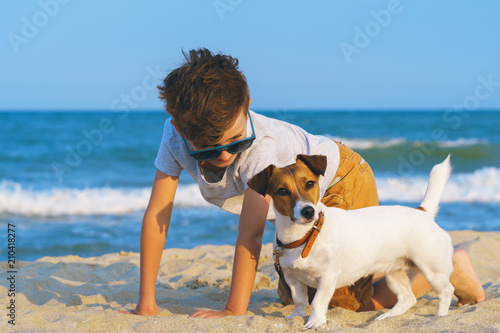 Fototapeta Naklejka Na Ścianę i Meble -  Happy boy hugging his dog breed Jack russell at the seashore against a blue sky close up at sunset. Best friends rest and have fun on vacation, play in the sand against the sea