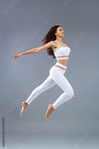 Attractive excited fitness girl, ballerina dancer in sportwear jumping of joy isolated over gray background.