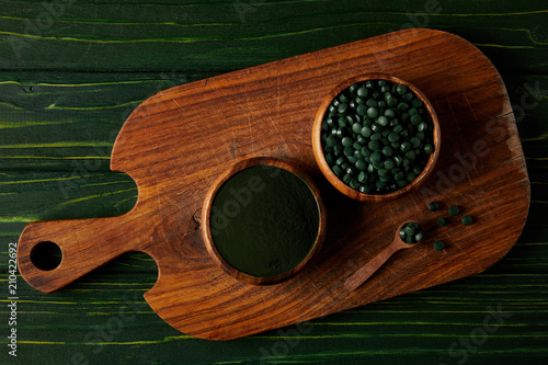 top view of cutting board with wooden spoon, bowls with spirulina powder and spirulina in pills on green wooden table photo