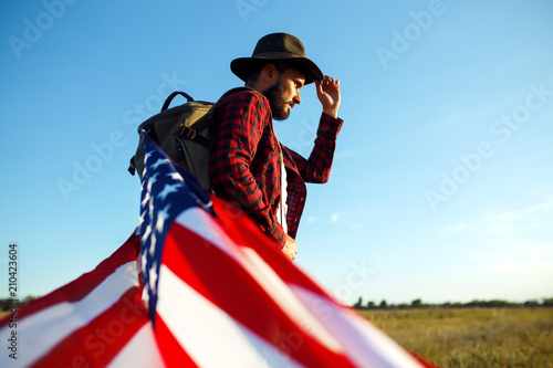 4th of July. American Flag.Patriotic holiday. Traveler with the flag of America. The man is wearing a hat, a backpack, a shirt and jeans. Beautiful sunset light. American style. 