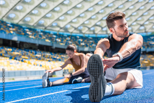 young athletic couple sitting on running track and stretching at sports stadium