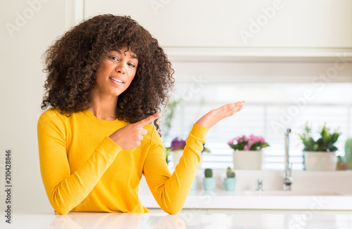 African american woman wearing yellow sweater at kitchen amazed and smiling to the camera while presenting with hand and pointing with finger.