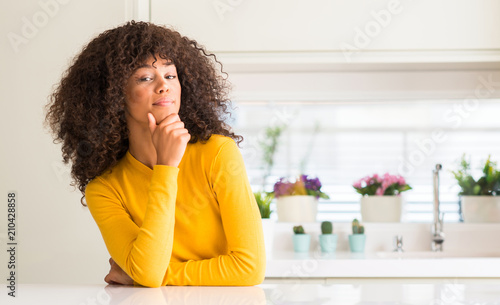 African american woman wearing yellow sweater at kitchen looking confident at the camera with smile with crossed arms and hand raised on chin. Thinking positive.