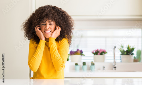 African american woman wearing yellow sweater at kitchen covering ears with fingers with annoyed expression for the noise of loud music. Deaf concept.