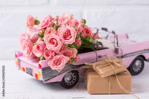Retro car toy with pink roses and wrapped boxes with presents  flowers against  white textured  wall.