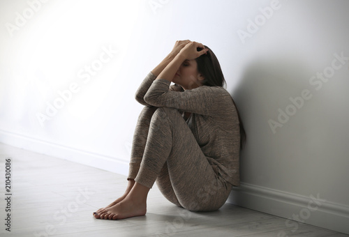 Depressed young woman sitting on floor near light wall