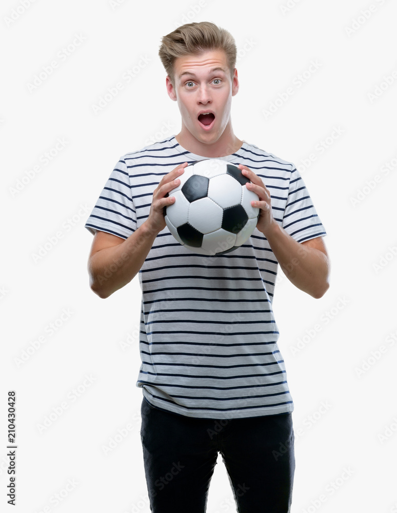 Young handsome blond man holding soccer ball scared in shock with a surprise face, afraid and excited with fear expression