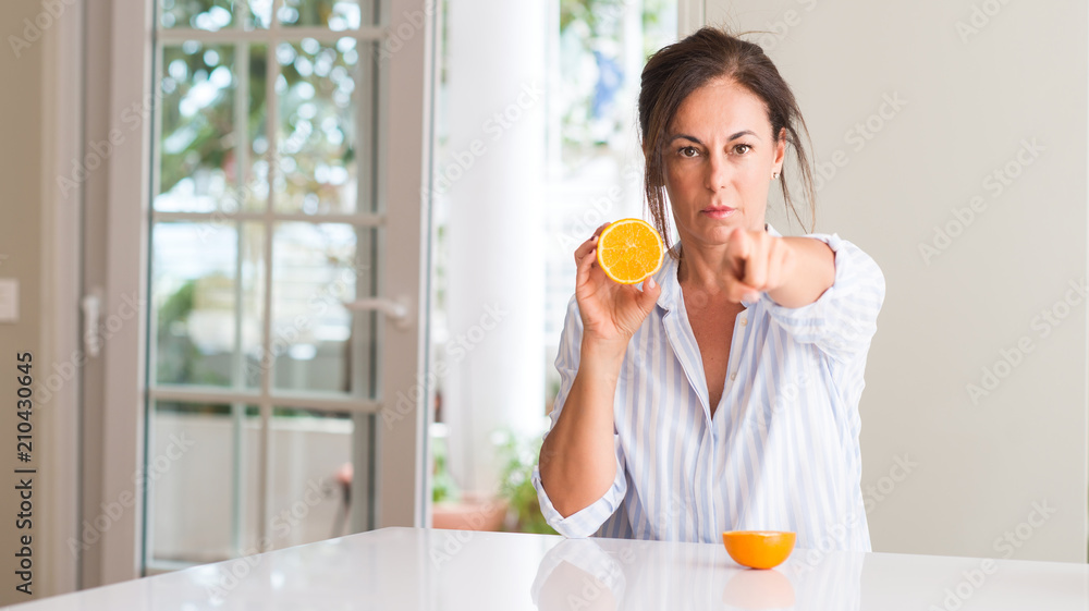 Middle aged woman holding orange fruit pointing with finger to the camera and to you, hand sign, positive and confident gesture from the front Middle aged woman holding orange fruit pointing with finger to the camera and to you, hand sign, positive and confident gesture from the front