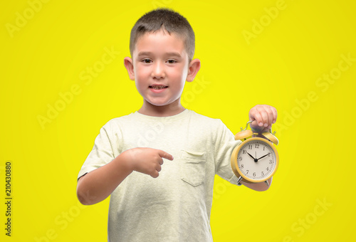 Little child holding alarm clock very happy pointing with hand and finger
