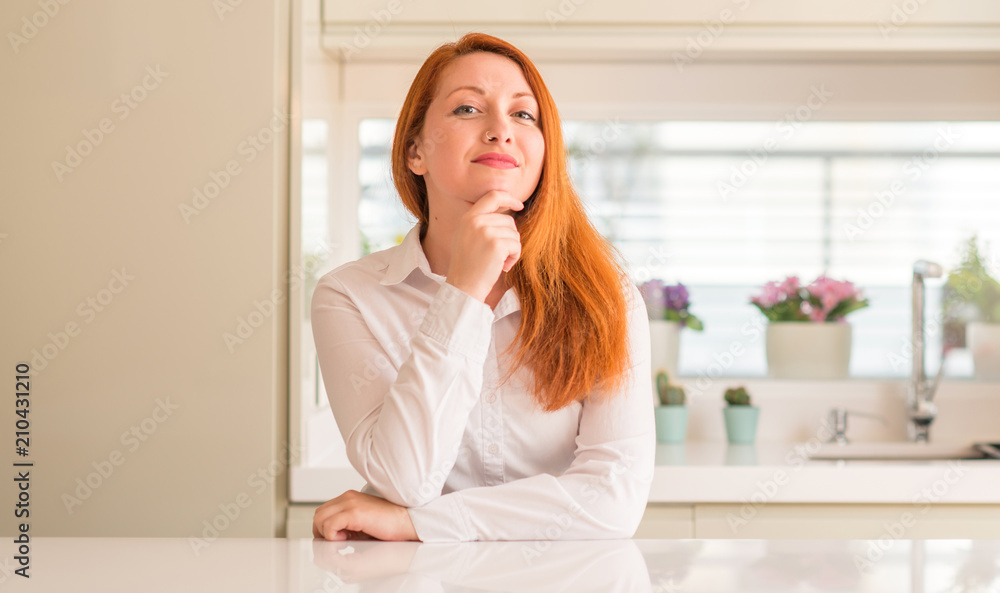 Redhead woman at kitchen looking confident at the camera with smile with crossed arms and hand raised on chin. Thinking positive.