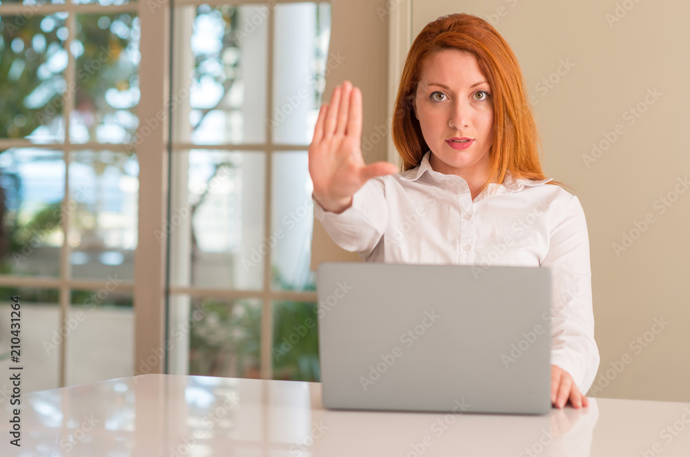 Redhead woman using computer laptop at home with open hand doing stop sign with serious and confident expression, defense gesture Redhead woman using computer laptop at home with open hand doing stop sign with serious and confident expression, defense gesture