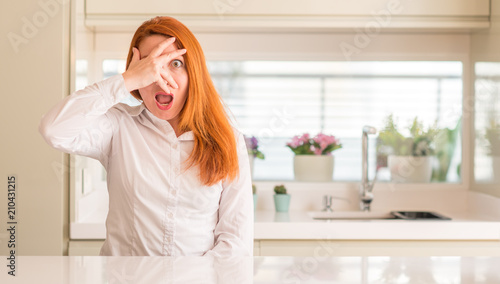 Redhead woman at kitchen peeking in shock covering face and eyes with hand  looking through fingers with embarrassed expression.