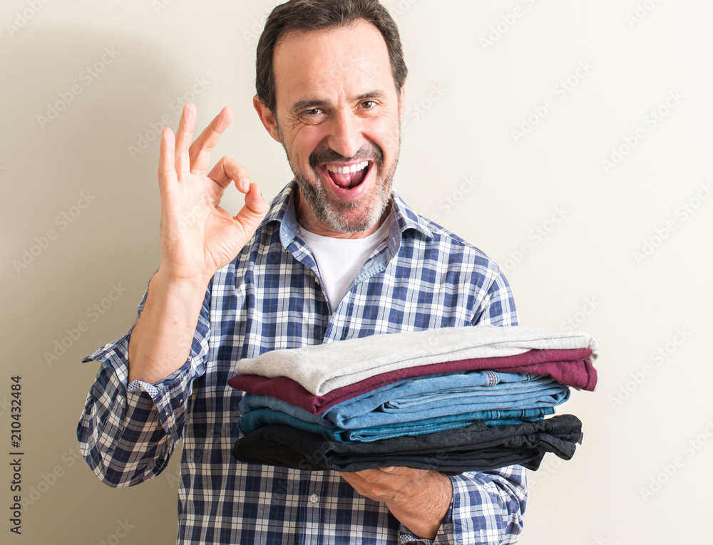 Senior man holding folded laundry clothes doing ok sign with fingers, excellent symbol Senior man holding folded laundry clothes doing ok sign with fingers, excellent symbol