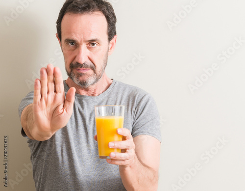 Senior man drinking orange juice in a glass with open hand doing stop sign with serious and confident expression, defense gesture