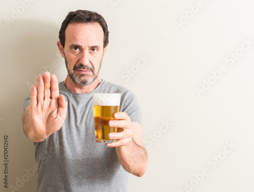 Senior man drinking beer with open hand doing stop sign with serious and confident expression, defense gesture