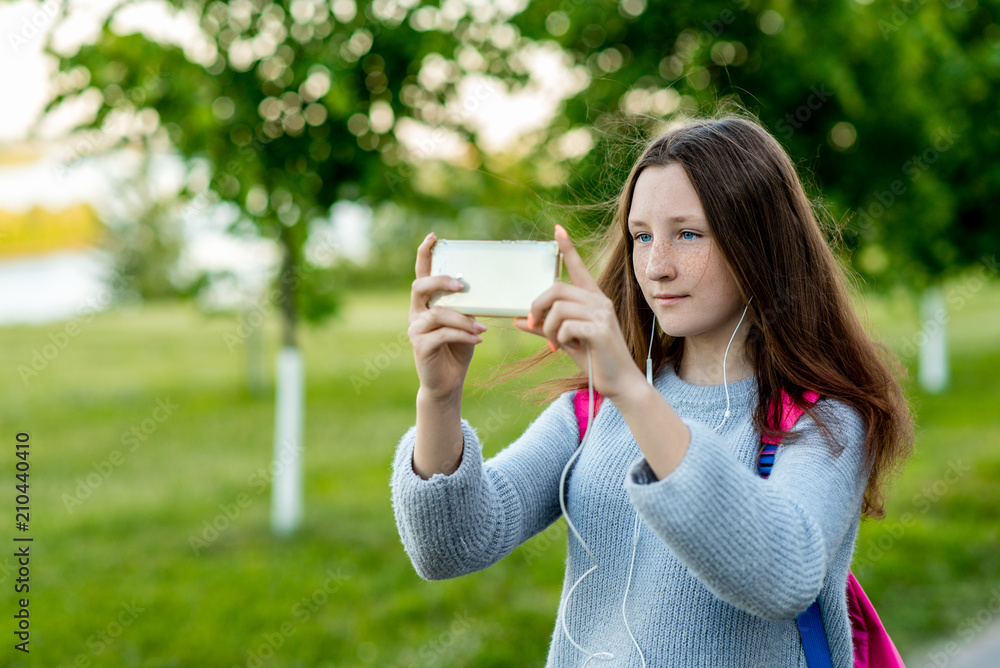 Beautiful schoolgirl girl. In the summer in park in nature. In his hands holds a smartphone. Take pictures of landscape. Free space for text. She smiles with freckles on her face. Rest after school. Beautiful schoolgirl girl. In the summer in park in nature. In his hands holds a smartphone. Take pictures of landscape. Free space for text. She smiles with freckles on her face. Rest after school.