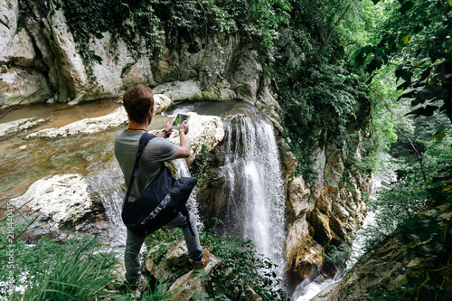 A traveler with a phone takes a picture of a large waterfall in the Agur gorge on the edge of a cliff